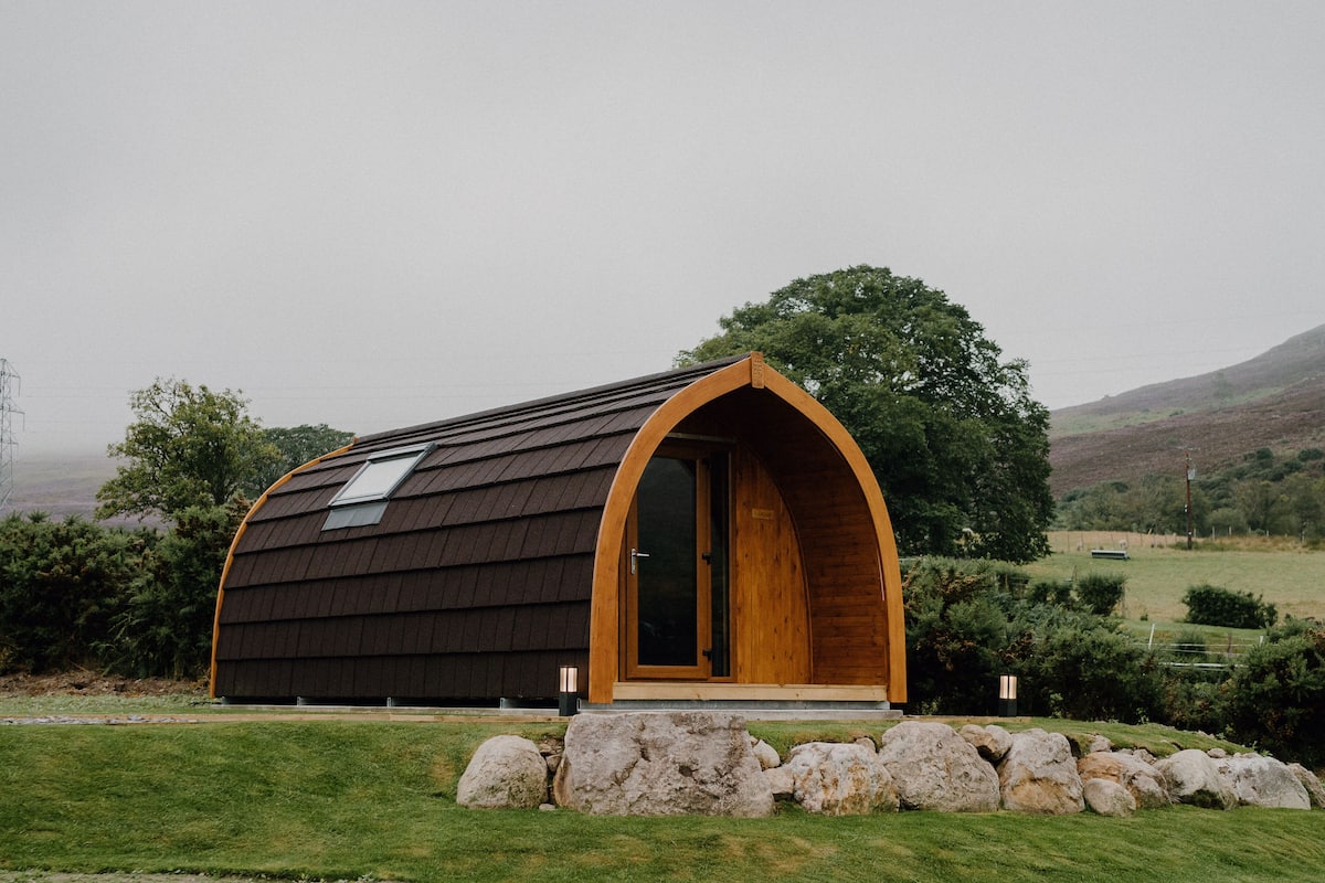 A small, modern, arched wooden structure with a sloped roof, surrounded by a grassy area and rocks, set in a rural landscape.