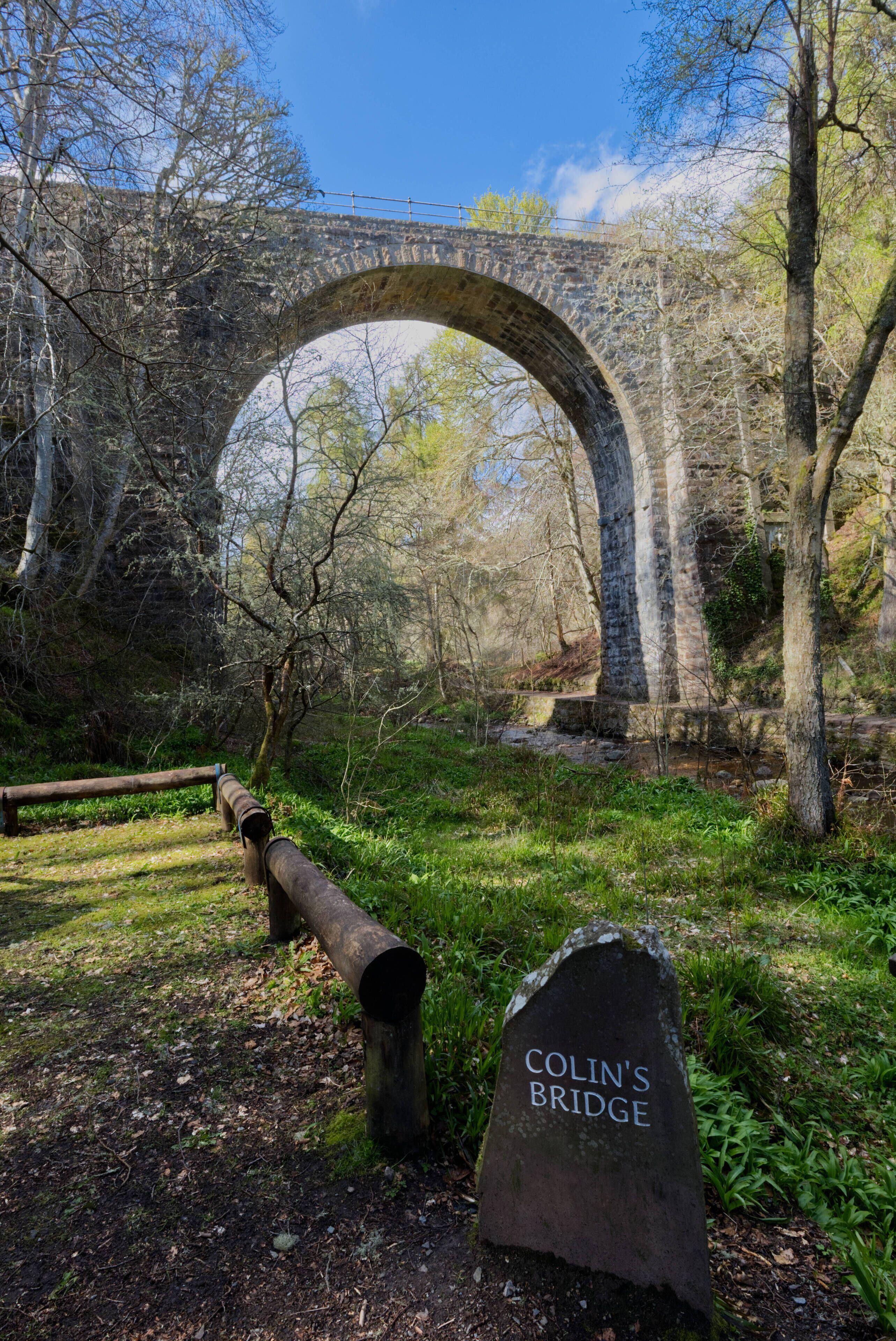 A stone bridge with a sign reading "Colin's Bridge" in a wooded area.