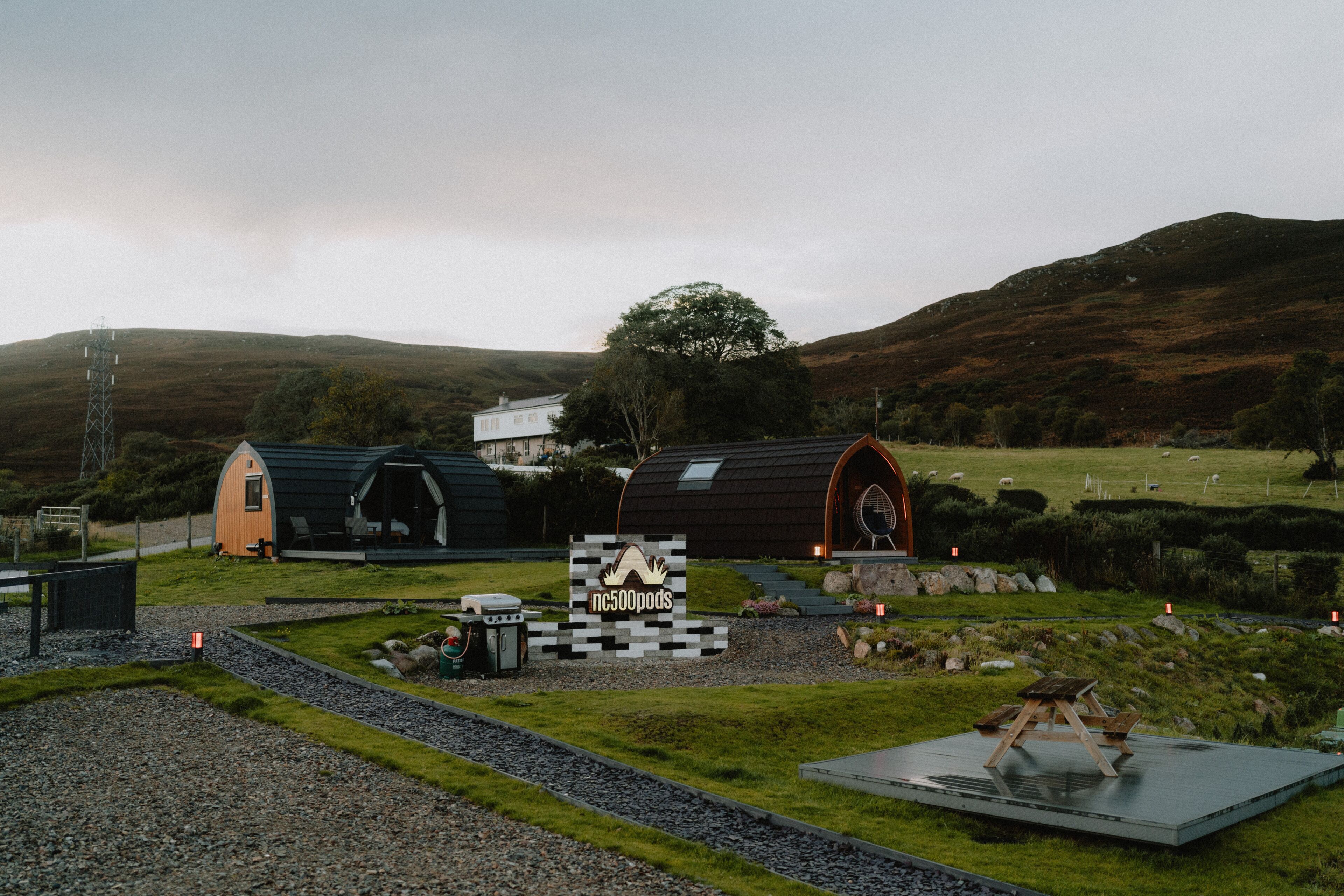 A rural accommodation with a distinctive black and white arch structure, a small wooden building, and a sign indicating 'nc500pods'.