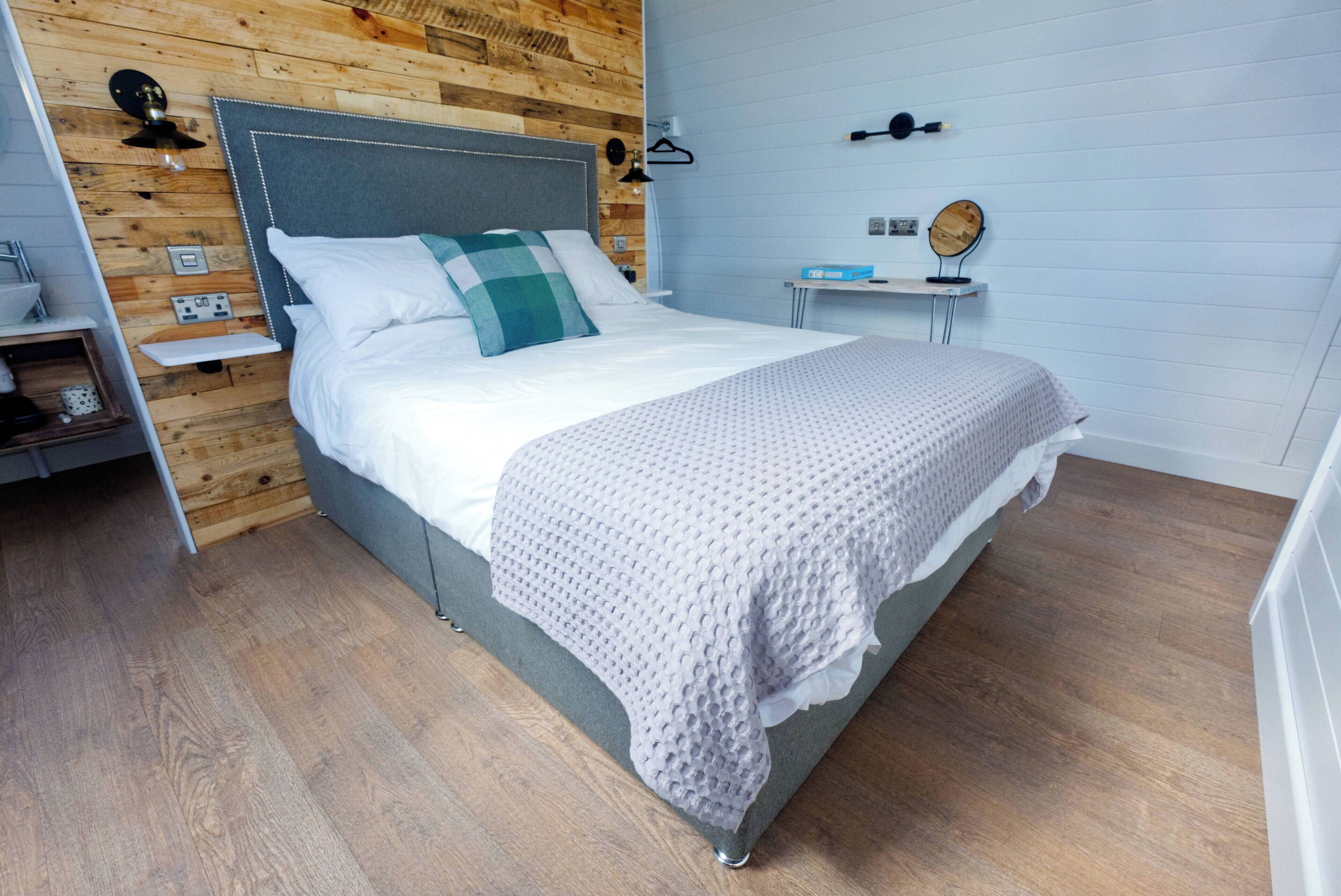 A modern bedroom with a wooden wall, a grey headboard, a white bed with a checkered pillow, a bedside table with a mirror, and a small shelf.