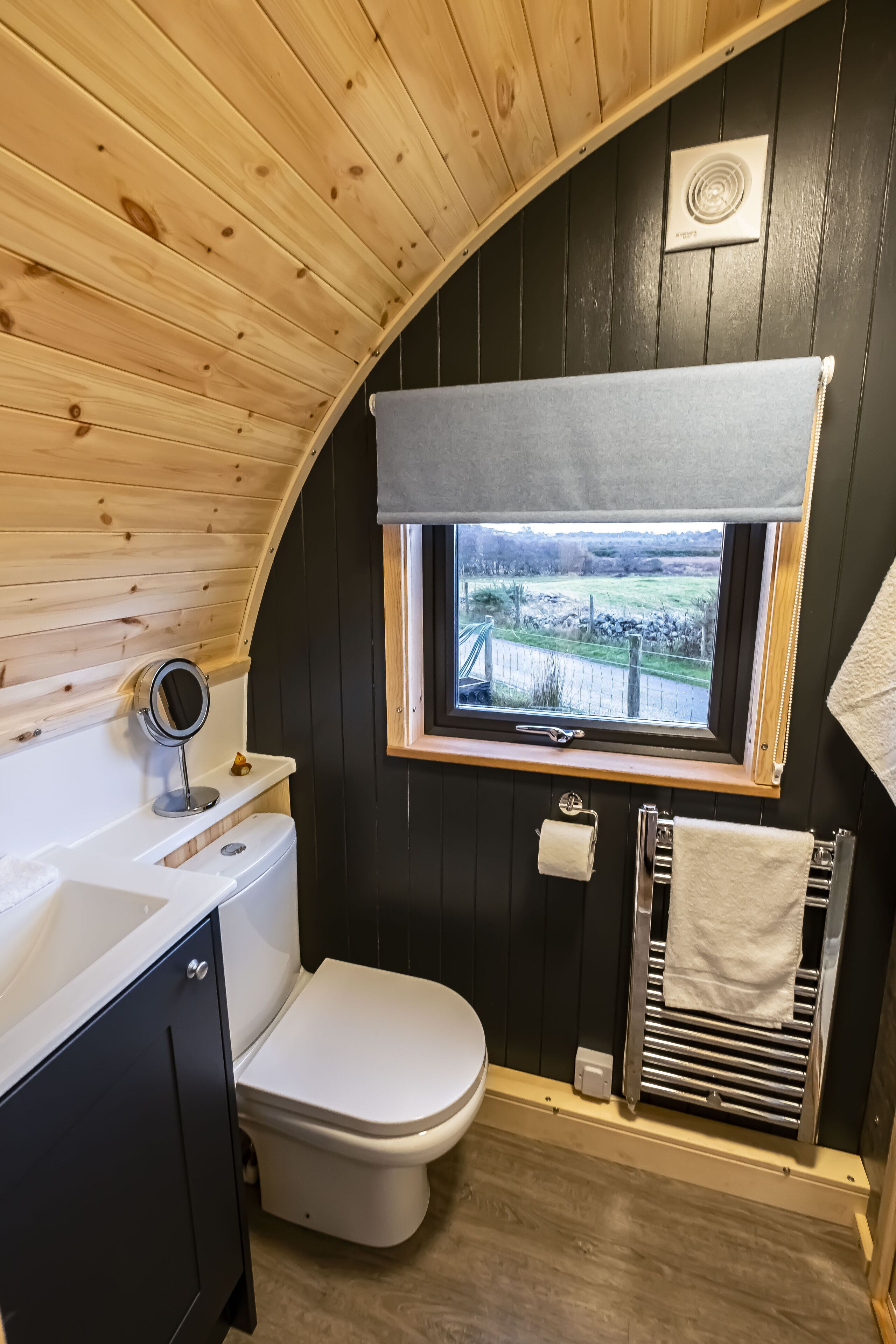 Un baño pequeño y curvo con techo de madera, ventana con vistas, inodoro, lavabo y toallero.