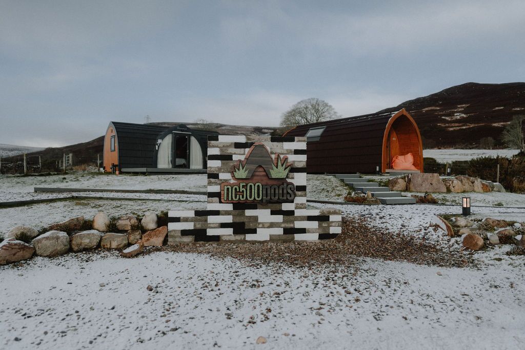 A sign with 'nc500pods' and a logo, surrounded by a stone wall and steps leading to pod-like structures.
