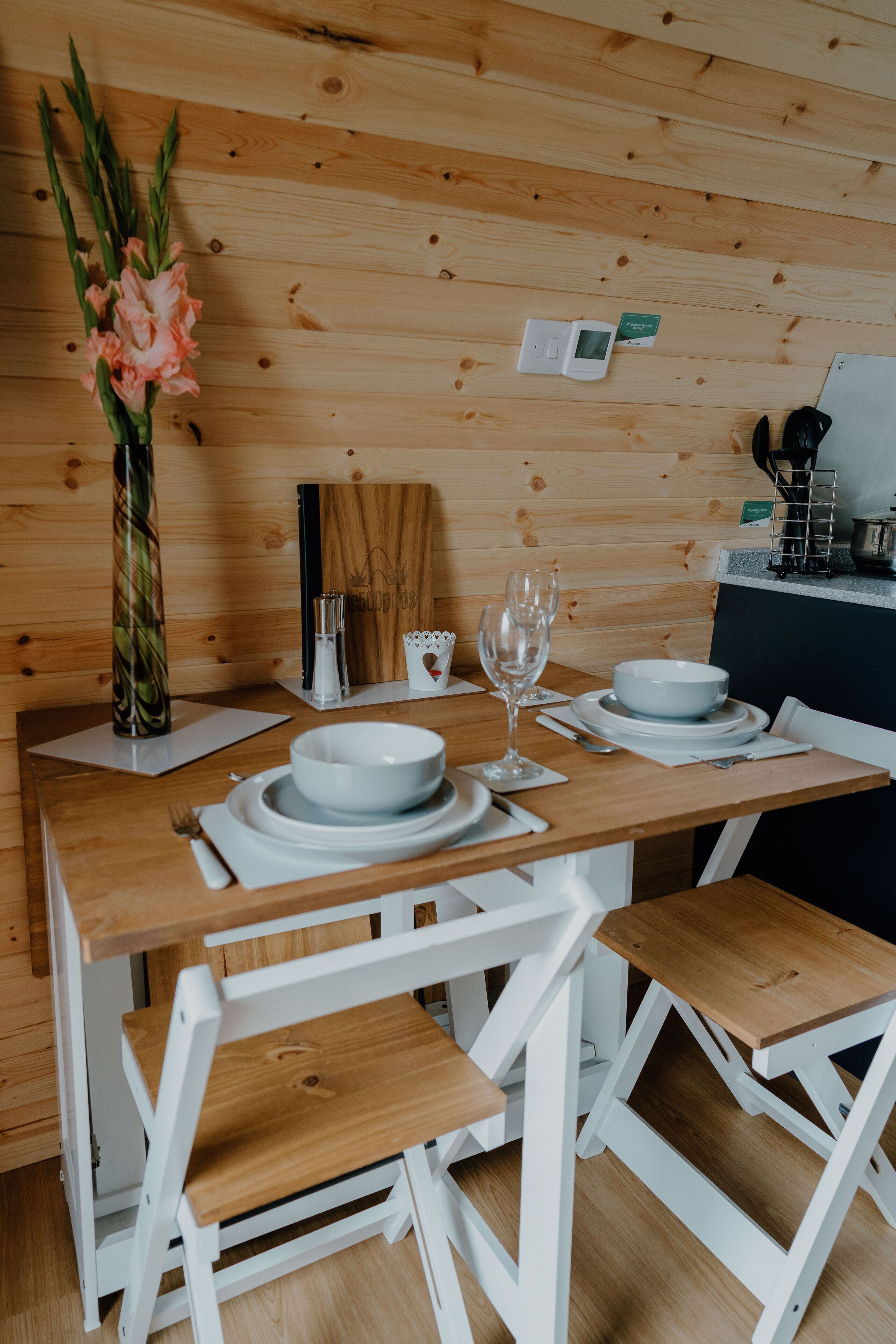 Ein Holztisch mit weißen Stühlen, eine Vase mit Blumen und ein digitaler Thermostat an der Wand.