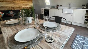 Un comedor con una mesa de madera dispuesta para cuatro personas, una cocina al fondo y un techo inclinado con tragaluces.