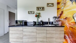 A modern kitchen with a central island, wooden cabinets, and a black countertop. There are framed pictures on the wall, a vase with flowers, and a coffee machine. - Lion King Suite - 4P near Disneyland Paris (Montry)