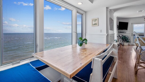 Ein Zimmer mit einem Holztisch und blauen, gepolsterten Sitzmöglichkeiten, großen Fenstern mit Meerblick und einer Treppe, die in einen oberen Bereich führt.