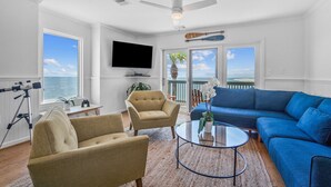 Ein Wohnzimmer mit einem blauen Sofa, zwei Sesseln, einem Glastisch, einem Deckenventilator, einem an der Wand montierten Fernseher und einem Teleskop auf einem Stativ.