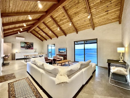 A spacious living area with a high ceiling, a large sofa, a dining table, and a view of the sea through the sliding glass doors.