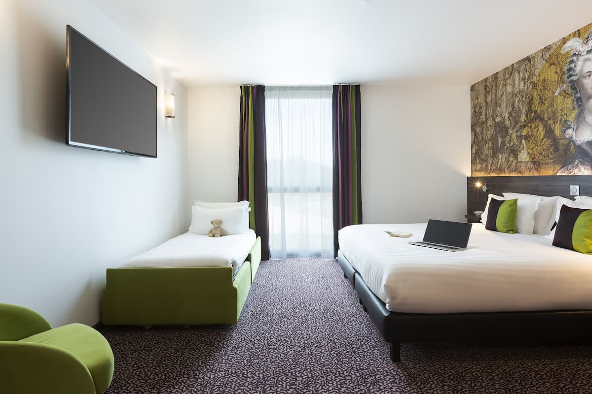 Une chambre d’hôtel avec un lit, un bureau avec un ordinateur portable, une chaise verte et un téléviseur fixé au mur.