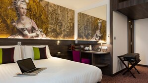 Habitación de hotel con una cama grande, una laptop sobre la cama, un escritorio con una silla, una mesita con una lámpara y un maletero sobre un carrito.