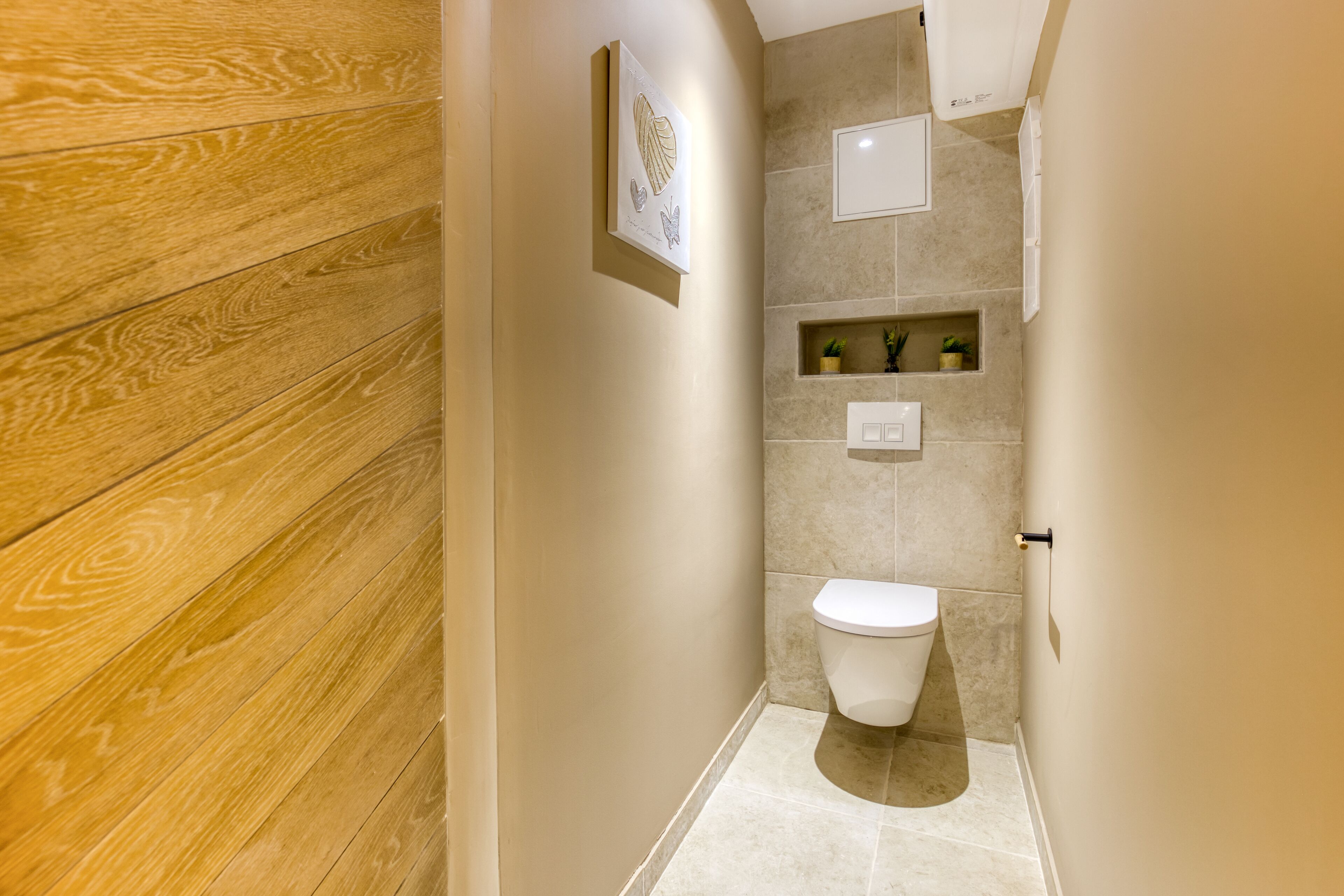 Un baño estrecho con piso de madera, inodoro blanco, una pequeña estantería con plantas y un cuadro enmarcado en la pared.