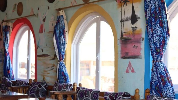 En restaurant med træborde og stole, store vinduer med gardiner og et farverigt mural på væggen.