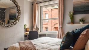 A bedroom with a bed, a mirror, a window with curtains, a desk, and a view of a brick building. - Stonesthrow Townhouse (Leek)