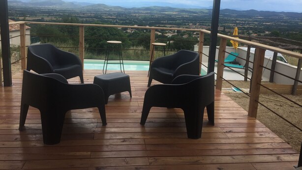 Een houten terras met twee zwarte stoelen en een kleine tafel, met uitzicht op een zwembad en verafgelegen bergen.