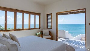 A bedroom with a large bed, a chair, and a view of the ocean through large windows.