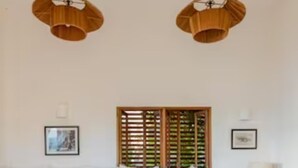 A modern living room with a large wooden coffee table, two wicker chairs with yellow cushions, a white sofa, a wooden slatted window, and two pendant lights.