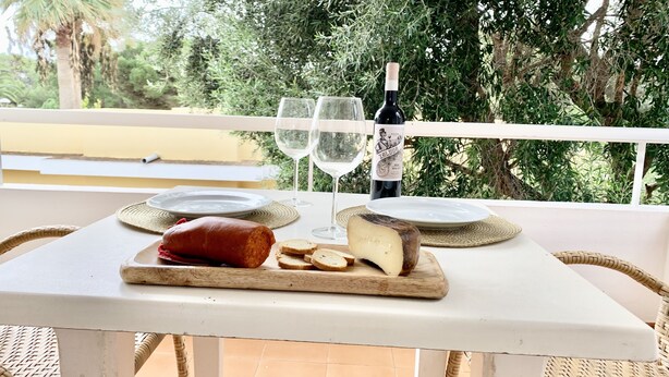 Una mesa con un pan, un queso redondo, dos vasos y una botella de vino en un balcón con vistas a árboles.