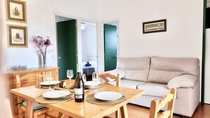 Un comedor con una mesa de madera dispuesta para dos personas, una botella de vino y un jarrón con flores. Se observa un sofá al fondo y una puerta que conduce a otra habitación.