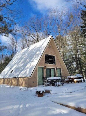 A cabin with a triangular roof, a dining table, and chairs outside.