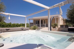 A hot tub area with a covered patio, a pool, and a building in the background. - VILLA NUCCI -ROSMARINO con jacuzzi esterna privata (Selva di Fasano)