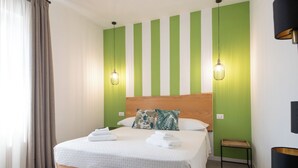 Een net opgemaakt bed met witte lakens, een houten hoofdbord en een gestreepte groene muur erachter. Aan beide zijden van het bed hangen twee wandlampen.