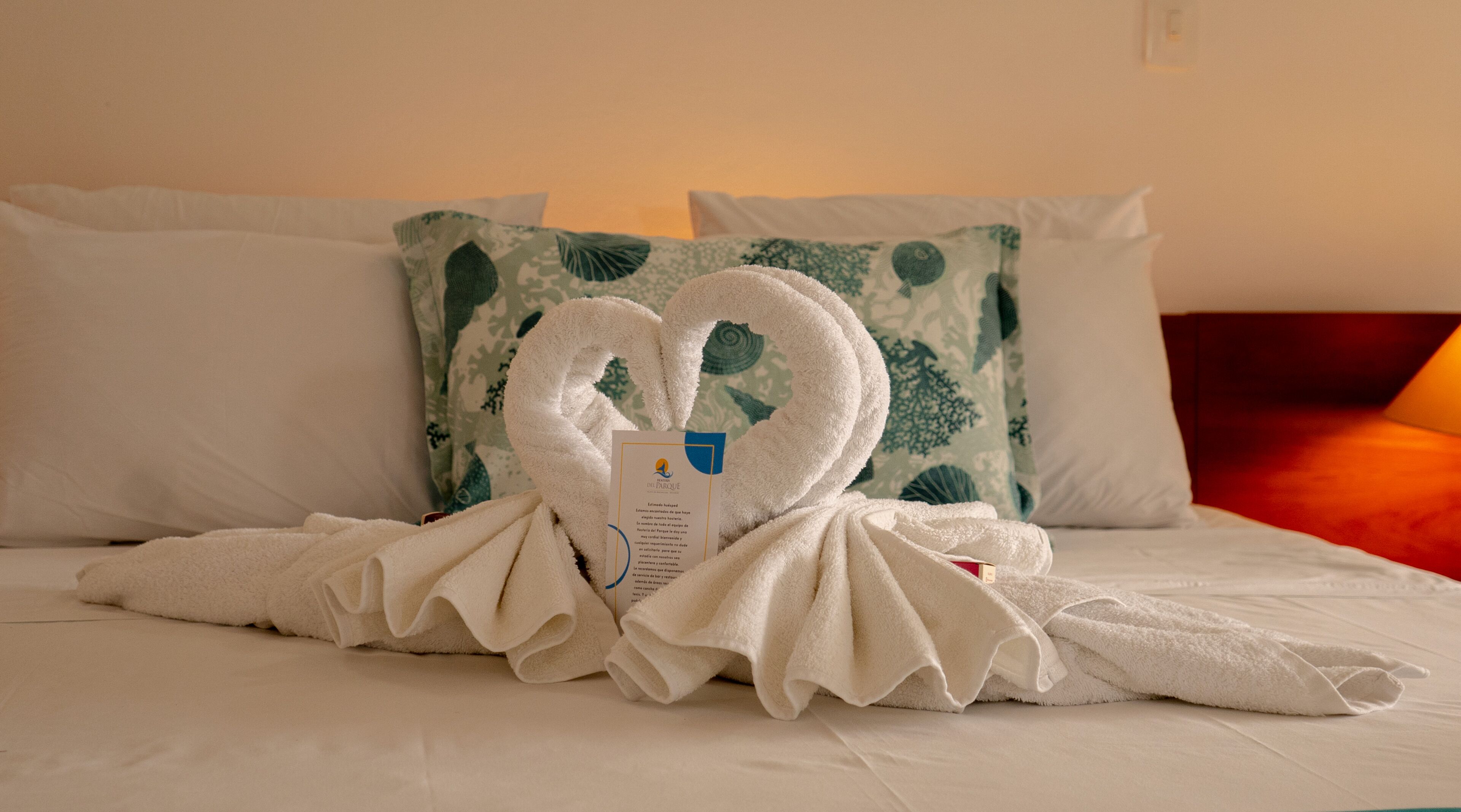 A hotel room with a neatly made bed, a heart-shaped towel arrangement, and a pillow with a leaf pattern.