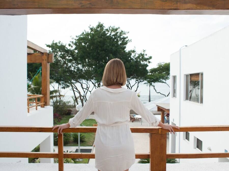 Una persona de pie en un balcón con vista a un edificio blanco y un árbol.