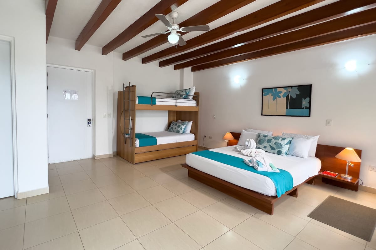 Quarto de hotel com beliche, uma cama de solteiro, ventilador de teto, porta e uma obra de arte na parede.