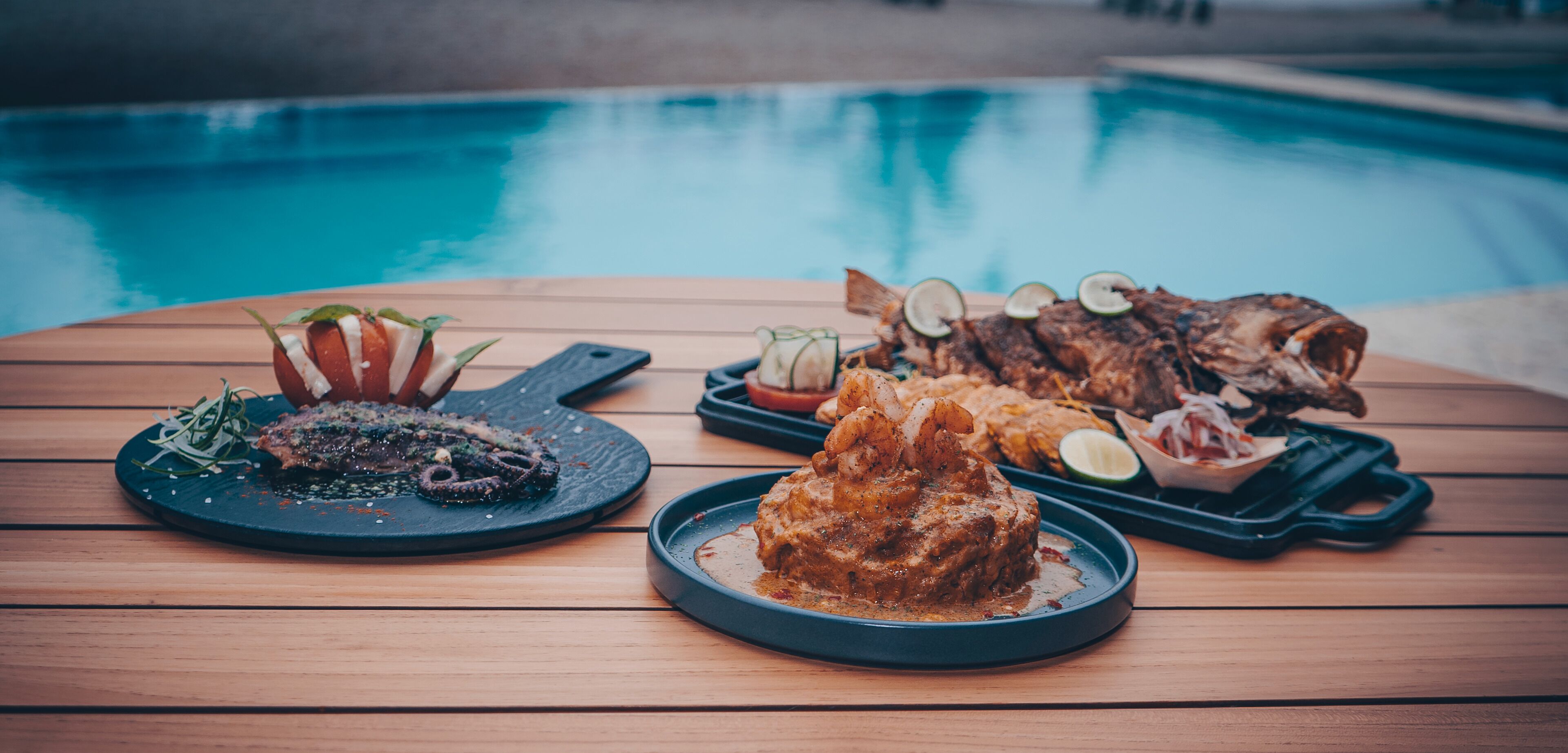 A wooden table with various dishes, including seafood and a steak, near a pool.