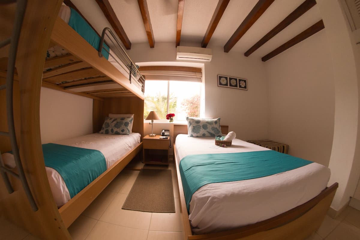 Quarto compacto com duas camas beliche, uma pequena mesa de cabeceira com luminária e janela com vista para o verde.