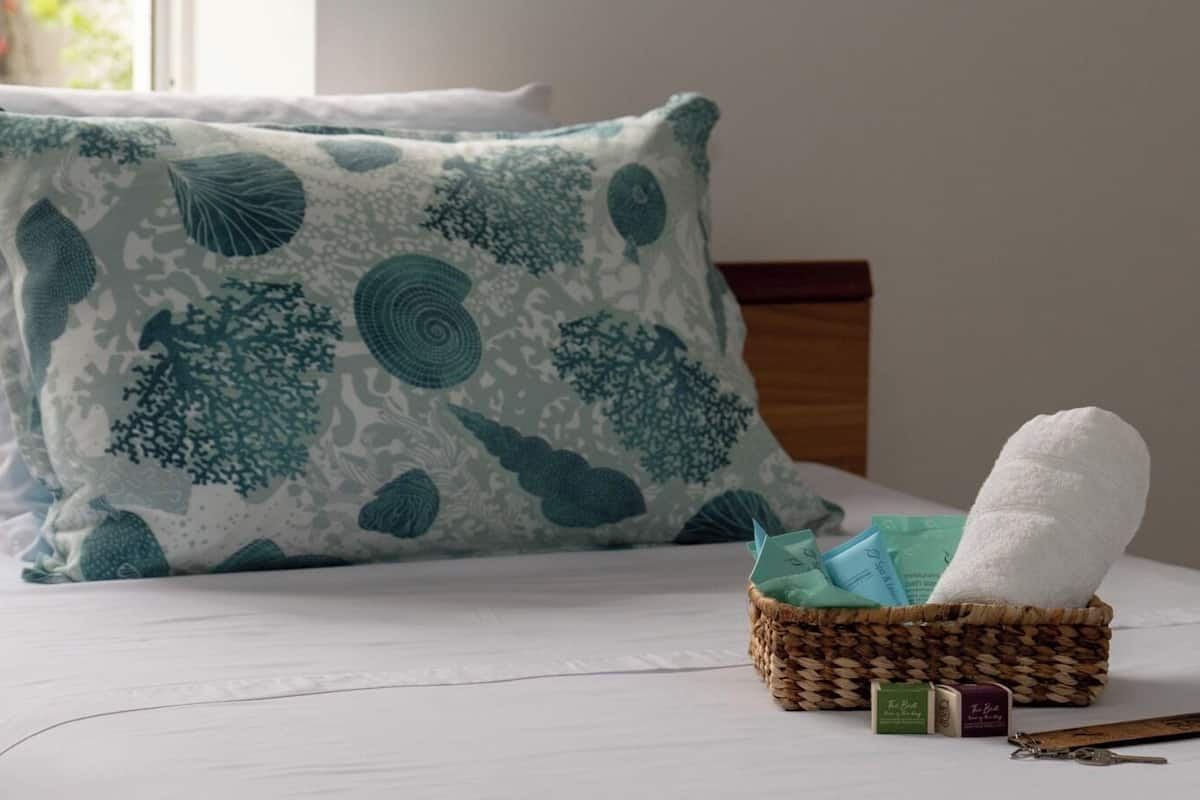 Uma cama com lençol branco, almofada estampada com folhas azul-esverdeadas e um cesto com toalha branca, artigos de higiene pessoal e uma chave.