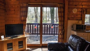 Ein Interieur einer Blockhütte mit Fernseher auf einem Holzschrank, einer Ledercouch, einer Schiebetür zu einer Terrasse und einer Mikrowelle auf einer Arbeitsplatte.