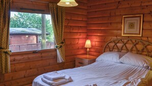 Ein Zimmer in einer Holzhütte mit einem Bett, einem Nachttisch, einer Lampe und einem Fenster mit Vorhängen.