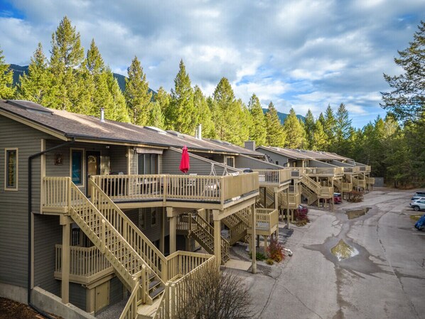 A row of cabins with wooden balconies and staircases, surrounded by trees and a parking area.