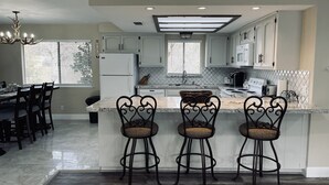 A modern kitchen with white cabinets, a central island with bar stools, and a dining area with a chandelier. -  Newly remodeled 3 bedroom home located near Yosemite and bass lake (Oakhurst)