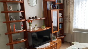 A living room with a TV, wooden shelves, a dining table, and a window with curtains. - Colorful city on the Elde 2 (Grabow)