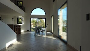 A spacious interior with a high ceiling, a dining area with chairs and a table, and large windows offering views of the outdoors.