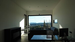 A modern living room with a large window, a sofa, a coffee table, and a flat-screen TV. - Secluded villa with heated pool and views of vineyards and sea (Figagnaire)