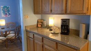 A kitchen with wooden cabinets, a granite countertop, a coffee maker, and a dining area with chairs and a table. - Mermaid's and Mutts Retreat - Beach Front, Pet Friendly, Wi-Fi, Off-Street Park (Norfolk)