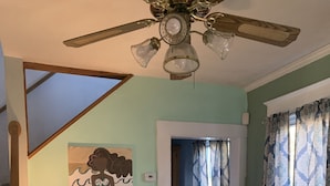 A dining area with a wooden table and chairs, a red table lamp, a ceiling fan, and a wall painting. - Mermaid's and Mutts Retreat - Beach Front, Pet Friendly, Wi-Fi, Off-Street Park (Norfolk)