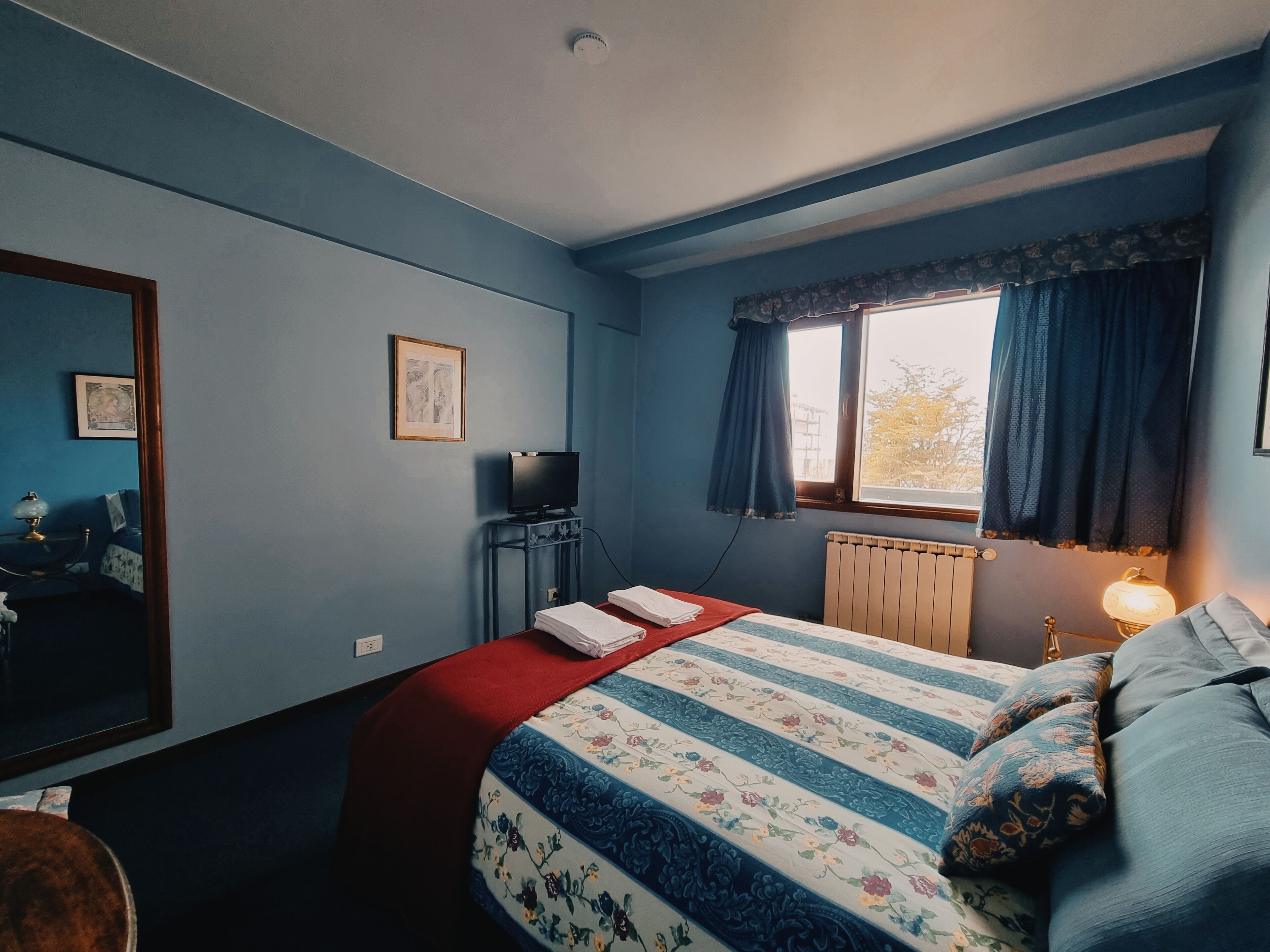 A bedroom with a bed, a TV, a window with curtains, a radiator, and a lamp.