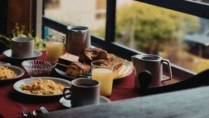 Une table de petit-déjeuner avec divers plats, un verre de jus d’orange et une tasse, vue à travers une fenêtre donnant sur une ville côtière.
