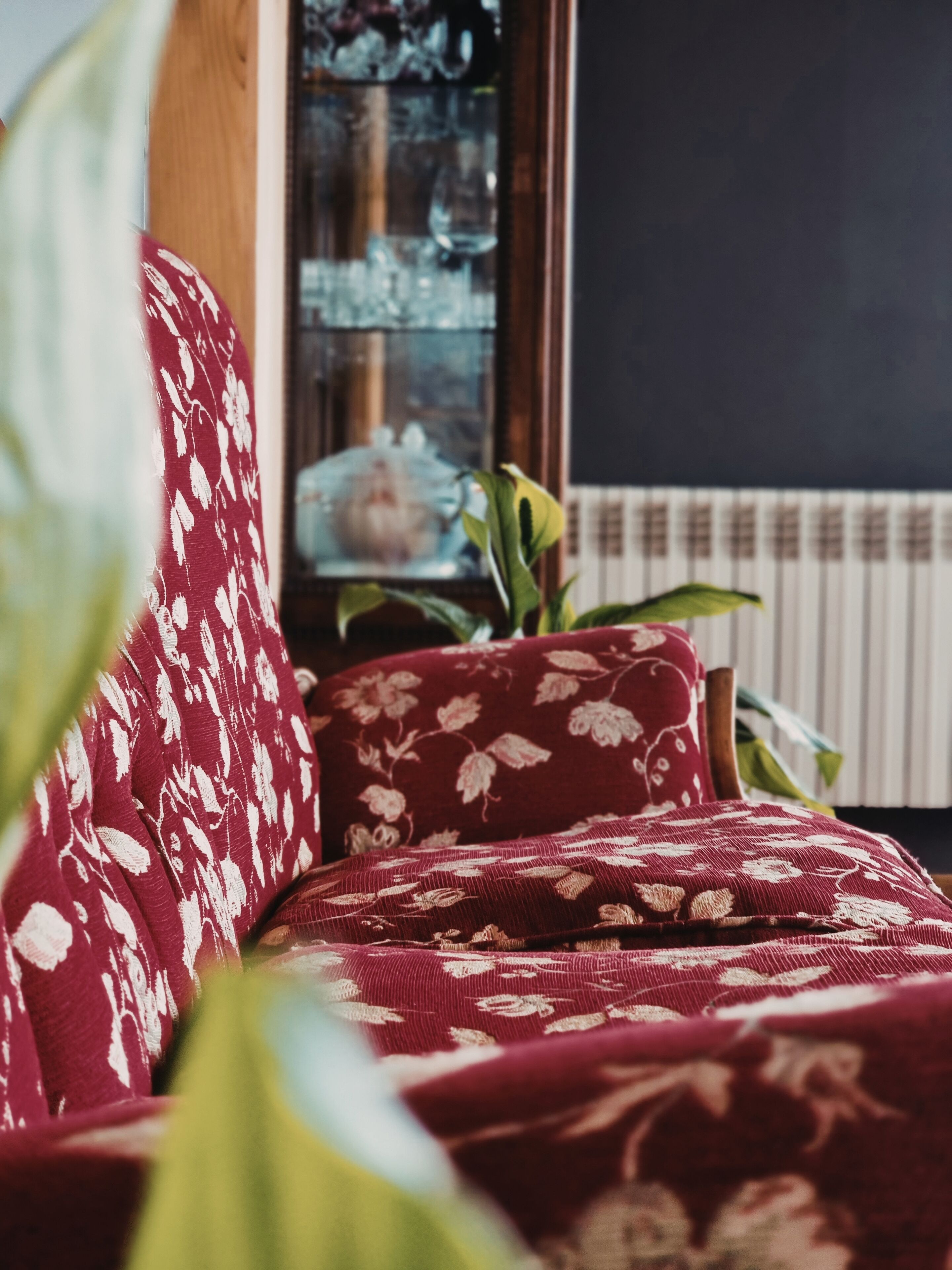 A red sofa with floral patterns, a wooden side table, and a potted plant in a room with a window and radiator.
