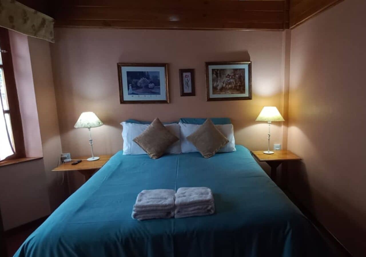 A neatly made bed with a teal bedspread, two pillows, and a stack of towels on the foot of the bed. There are two bedside lamps on wooden nightstands, and two framed pictures on the wall above the bed.