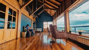 Un intérieur spacieux en bois, avec un plafond haut, de grandes fenêtres offrant une vue sur la mer, et un coin salon confortable avec un fauteuil et un canapé.