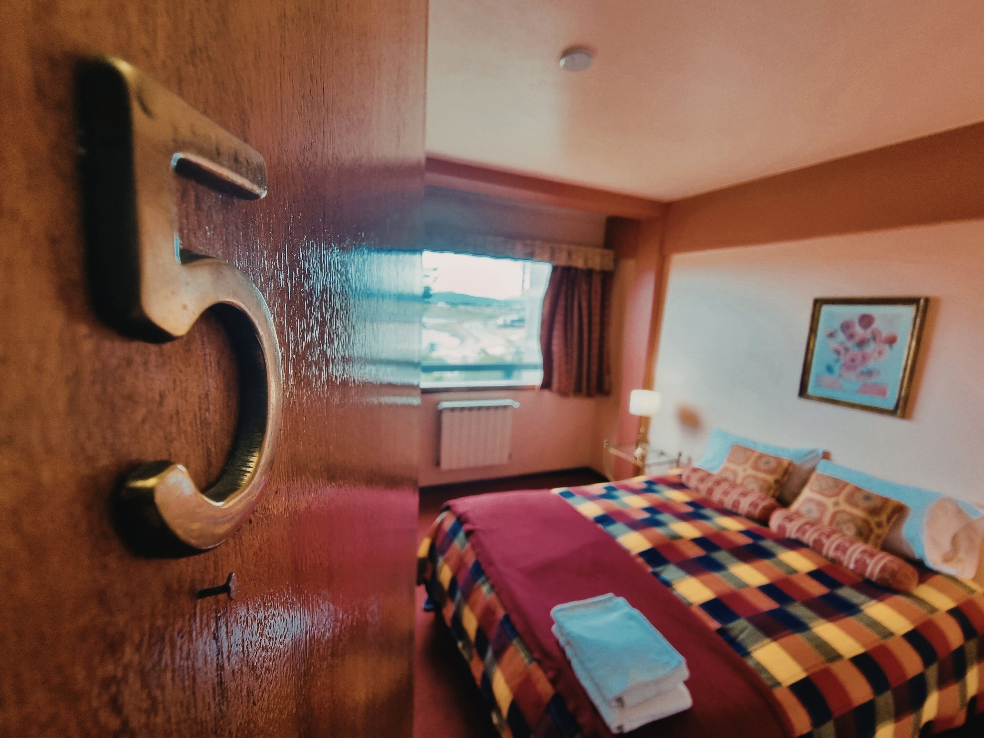 A hotel room with a wooden door marked with the number 5, a bed with a colorful checkered bedspread, a window with curtains, and a wall-mounted picture.