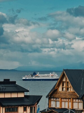 A cruise ship in the distance, a traditional wooden building in the foreground, and a body of water. - Flor Austral (Ushuaia)