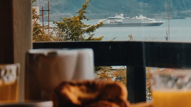 Une vue depuis une fenêtre donnant sur une étendue d’eau, avec un navire de croisière, des montagnes enneigées et un poteau téléphonique au loin.