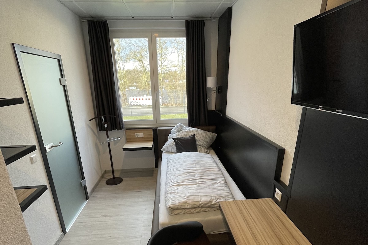 A compact hotel room with a single bed, a desk, a chair, a TV, and a window with curtains.