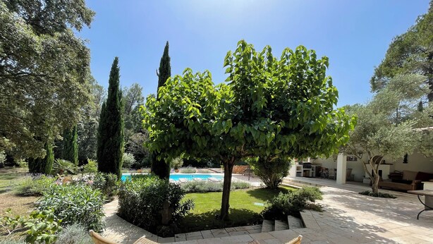 Un patio al aire libre con mobiliario de mimbre, una piscina y varios árboles y arbustos.